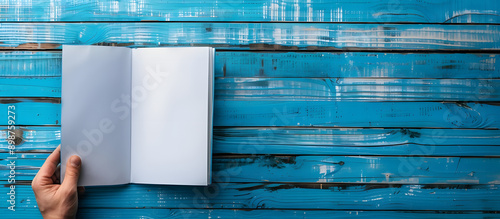 Looking down on a blue wooden surface, a man's hand holds a blank booklet, creating a minimalist composition with copy space image.