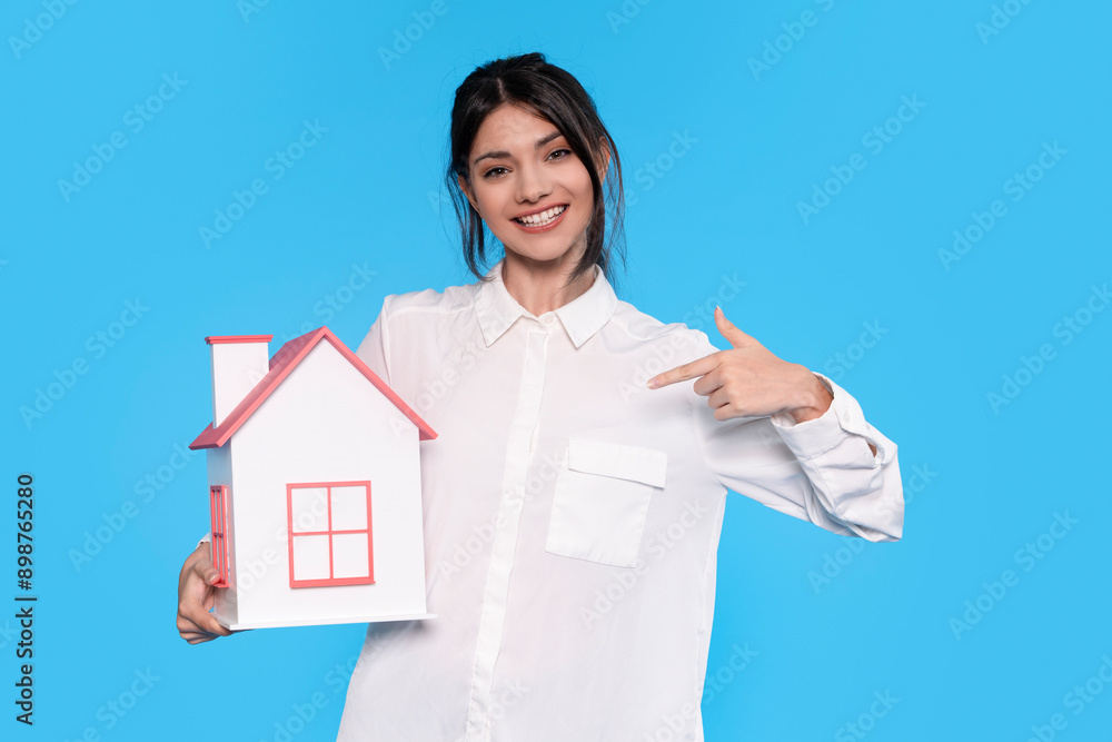 Woman in White Shirt Holding Miniature House and Pointing Towards It Against Blue Background Woman in White Shirt Holding Miniature House and Pointing Towards It Against Blue Background