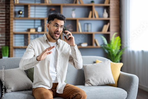 Angry young hispanic man sitting on sofa at home and aggressively talking on mobile phone