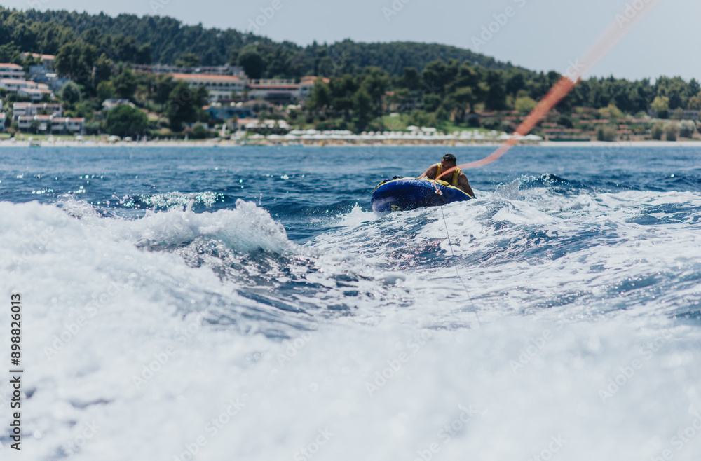 Naklejka premium Man enjoys an adventurous summer activity of tubing behind a speedboat on the sea, with a scenic coastal background.