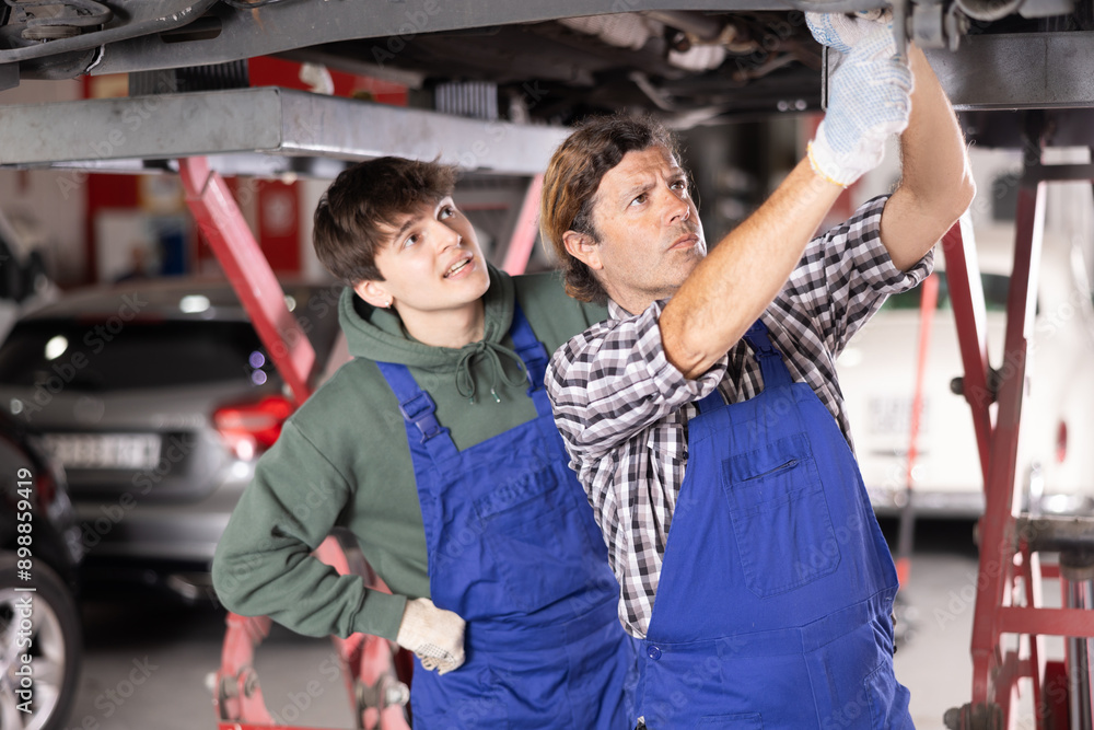 Obraz premium Young auto mechanic helps an experienced auto mechanic repair a car in a car service center