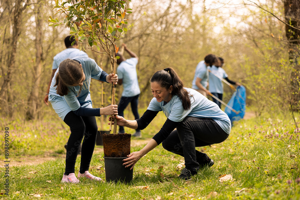 Fototapeta premium Mom and her little girl taking a tree out of the pot and plant it, doing voluntary work to help with reforestation and environmental protection. Activists taking action to help natural habitat.