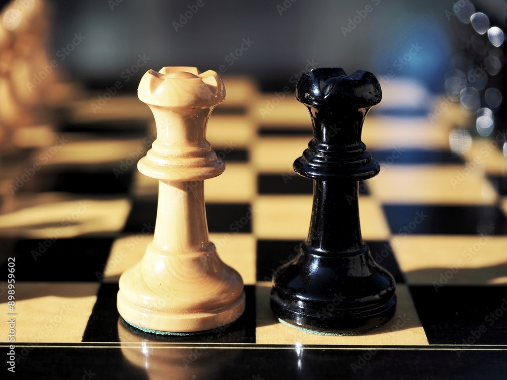 A close-up of two wooden black and white queen chess pieces isolated on a chessboard A close-up of two wooden black and white queen chess pieces isolated on a chessboard