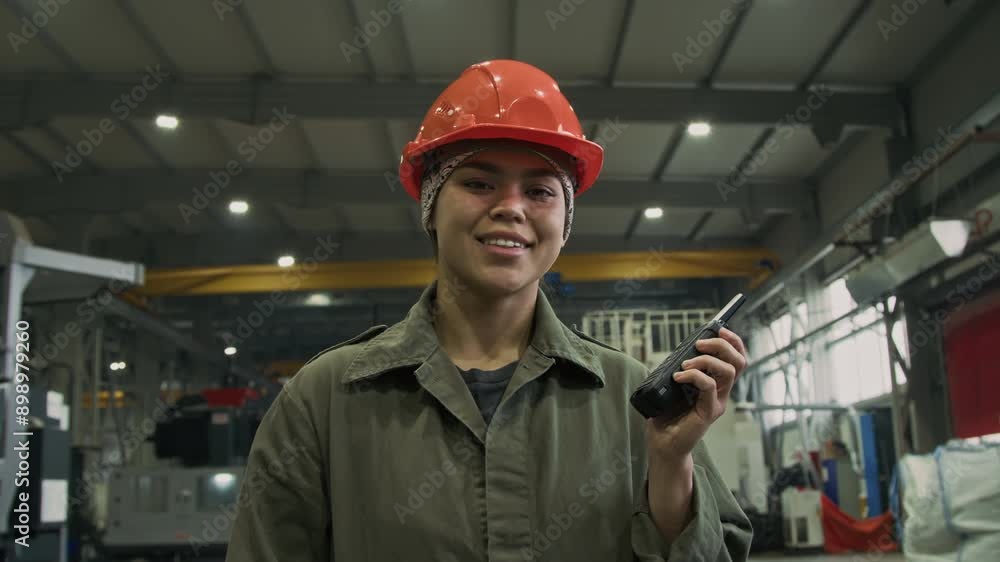 Chest up slowmo portrait of young Hispanic forewoman in orange hardhat and coveralls posing in factory workshop with walkie-talkie in hand, looking at camera Chest up slowmo portrait of young Hispanic forewoman in orange hardhat and coveralls posing in factory workshop with walkie-talkie in hand, looking at camera