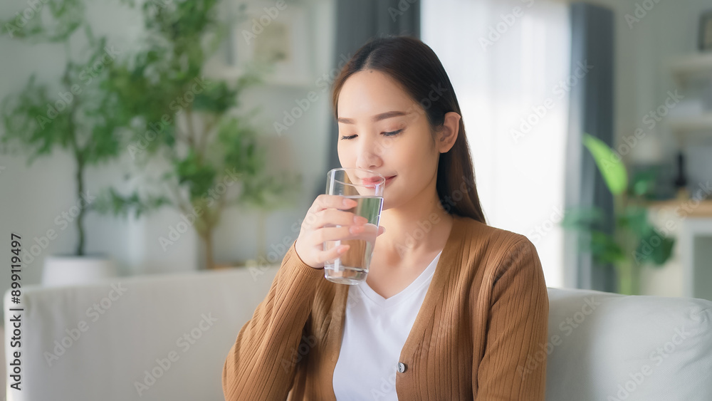 Happy young Asian woman drinking water while sitting on the sofa in the living room Happy young Asian woman drinking water while sitting on the sofa in the living room