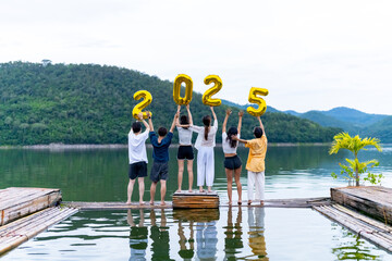 Group of Young Asian man and woman friends celebration happy new year party holding golden number 2025 balloon. People enjoy and fun travel nature mountain together at lake house on holiday vacation.