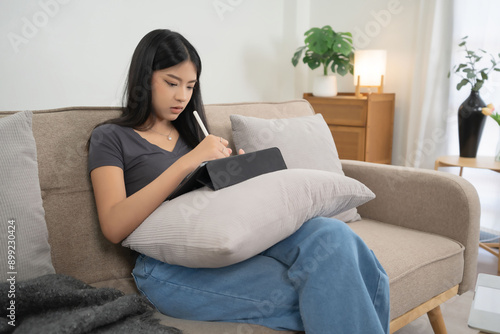 Young asian women reading business report about marketing and finance data of project and taking notes in tablet while sitting on couch to spending time and relaxation with enjoying lifestyle at home