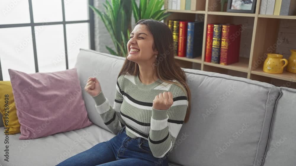 Victory at home! excited young hispanic woman, mad-crazy with success, celebrating her win sitting on sofa, arms raised in triumph Victory at home! excited young hispanic woman, mad-crazy with success, celebrating her win sitting on sofa, arms raised in triumph
