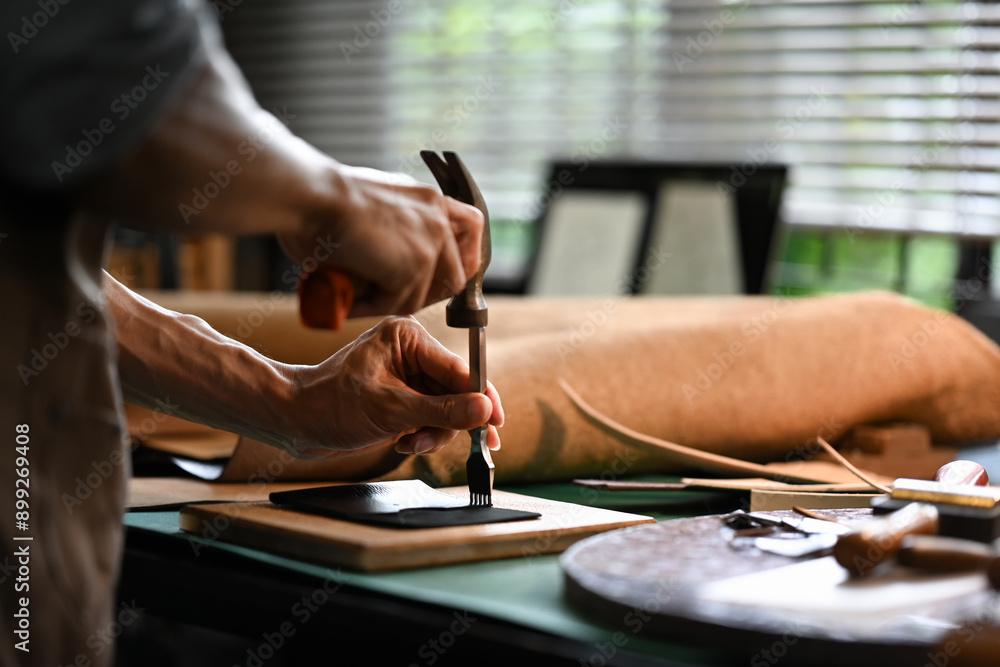 Obraz premium Asian male artisan carefully punching piece of leather at professional leather workshop