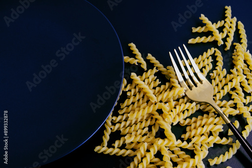 Fusilli pasta on black background. Top view of fusilli pasta, black plate, and fork with copy space. Concept of Italian food, raw ingredients, cooking, kitchenware. Close-up raw fusilli pasta texture.