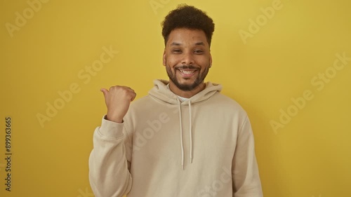 Cheerful african american man wearing a hoodie, flashes a broad smile while pointing to the side with thumb up - looking absolutely chuffed on an isolated yellow background