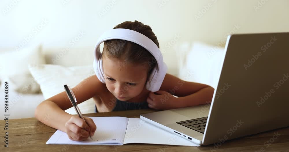 Focused little primary school pupil girl in headphones studying on Internet, writing learning notes in copybook, watching educational lesson, video class on laptop computer
