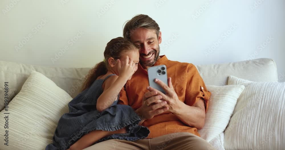 Cheerful Hispanic dad and cute girl taking family selfie on mobile phone at home, resting and hugging on couch, holding smartphone, posing, grimacing, laughing, using funny multimedia app for picture Cheerful Hispanic dad and cute girl taking family selfie on mobile phone at home, resting and hugging on couch, holding smartphone, posing, grimacing, laughing, using funny multimedia app for picture