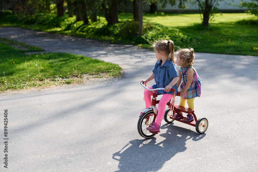 Little sisters ride a red tricycle together down the street. Little sisters ride a red tricycle together down the street.
