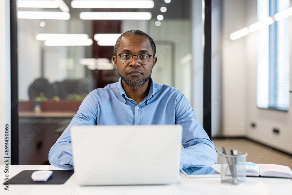 Focused professional working on laptop in modern office environment. Man wearing glasses, blue shirt, sitting at desk with notebook, mouse, and pen holder. Bright office with modern lighting. Focused professional working on laptop in modern office environment. Man wearing glasses, blue shirt, sitting at desk with notebook, mouse, and pen holder. Bright office with modern lighting.