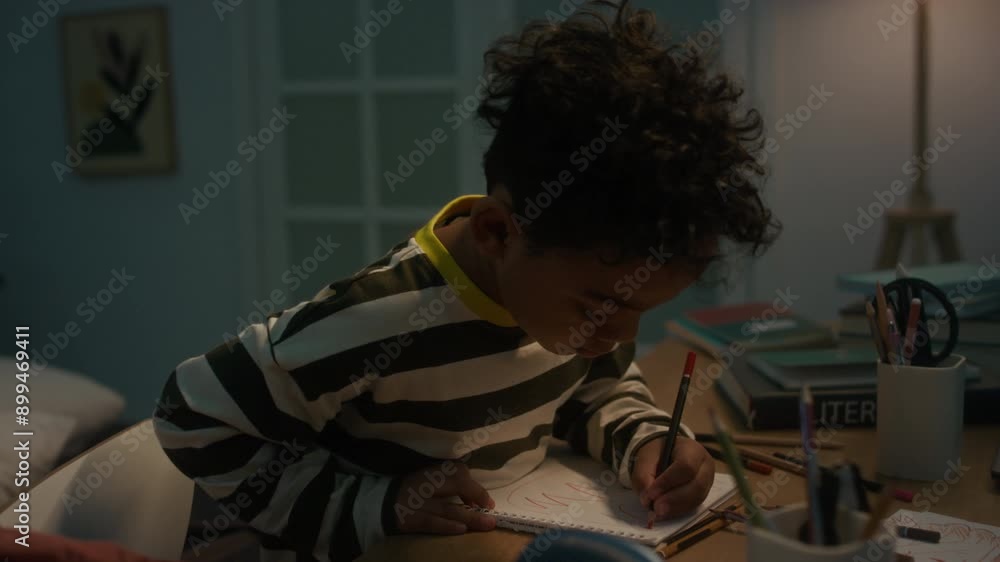 Medium shot of little African American boy drawing with pencil in notebook at home table Medium shot of little African American boy drawing with pencil in notebook at home table