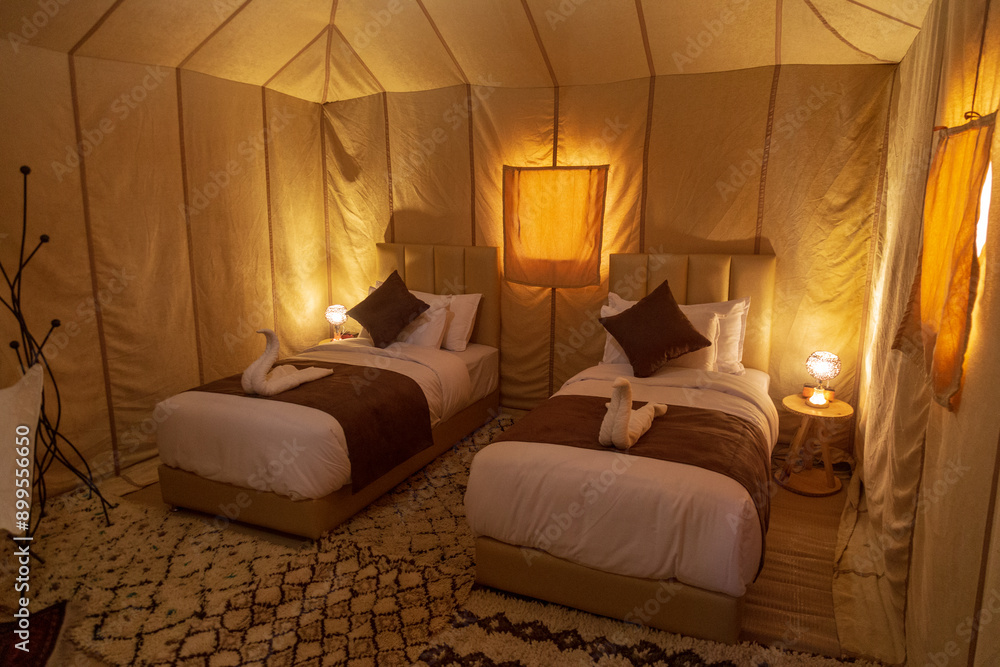 Moroccan attire walks through a desert camp at dusk, with white tents and lanterns arranged on colorful rugs, evoking a sense of cultural richness and hospitality. Moroccan attire walks through a desert camp at dusk, with white tents and lanterns arranged on colorful rugs, evoking a sense of cultural richness and hospitality.