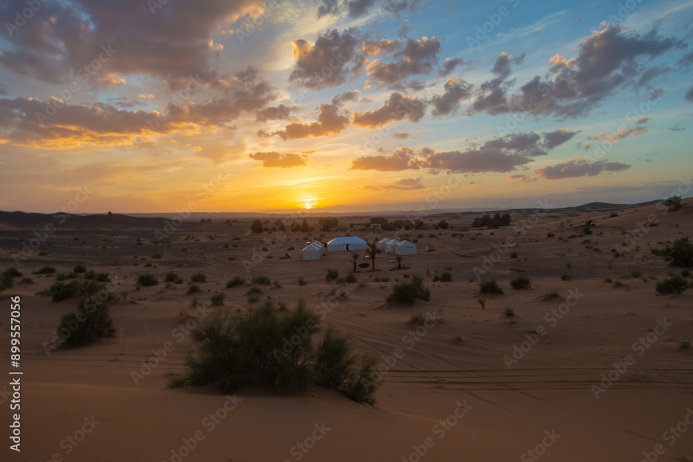 Moroccan attire walks through a desert camp at dusk, with white tents and lanterns arranged on colorful rugs, evoking a sense of cultural richness and hospitality. Moroccan attire walks through a desert camp at dusk, with white tents and lanterns arranged on colorful rugs, evoking a sense of cultural richness and hospitality.