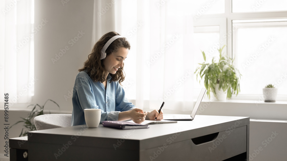 Focused young woman in headphones studying at home using laptop. Student girl e-learning sit at desk writing notes, jotting helpful information in copybook, makes high-school assignment, do homework Focused young woman in headphones studying at home using laptop. Student girl e-learning sit at desk writing notes, jotting helpful information in copybook, makes high-school assignment, do homework