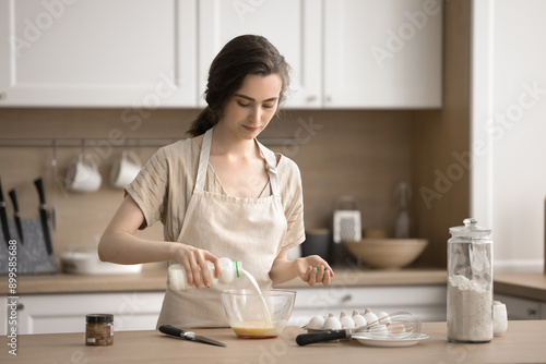 Young female pouring milk into bowl of beaten eggs, prepare omelet or dough for pastries, making homemade dish, enjoy process and homemade cookery process, trying new recipe. Routine, household chores © fizkes