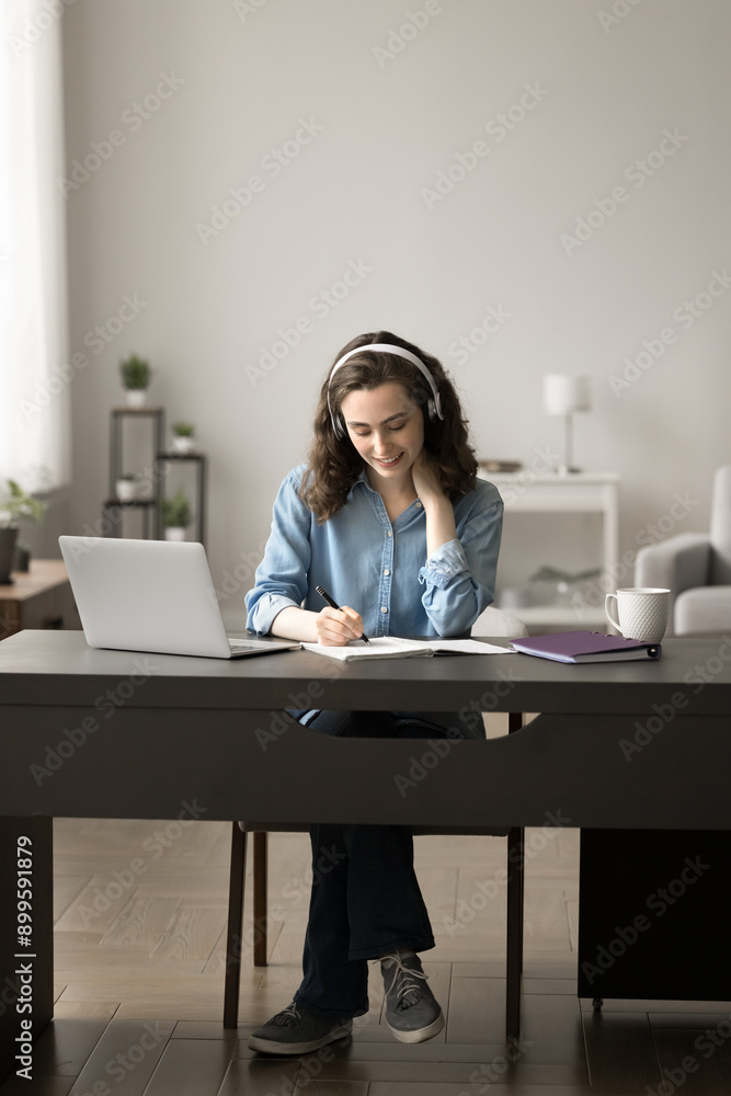 Attentive student girl in headphone engaged in on-line studying process, e-learning sit at desk with modern laptop, writing assignment in copybook, looks focused and interested to gain new knowledge Attentive student girl in headphone engaged in on-line studying process, e-learning sit at desk with modern laptop, writing assignment in copybook, looks focused and interested to gain new knowledge