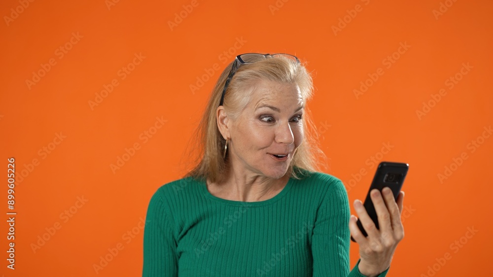 Smiling excited success mature woman posing isolated on orange background in studio. People sincere emotions, lifestyle concept. Pointing to cellphone using mobile phone happy win Smiling excited success mature woman posing isolated on orange background in studio. People sincere emotions, lifestyle concept. Pointing to cellphone using mobile phone happy win