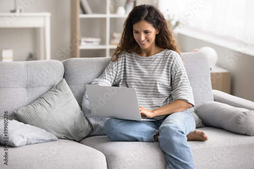 Woman sit on sofa with modern laptop, browsing wireless internet, enjoy on-line shopping, reading news, working or studying remotely, researching information, participating in online courses at home