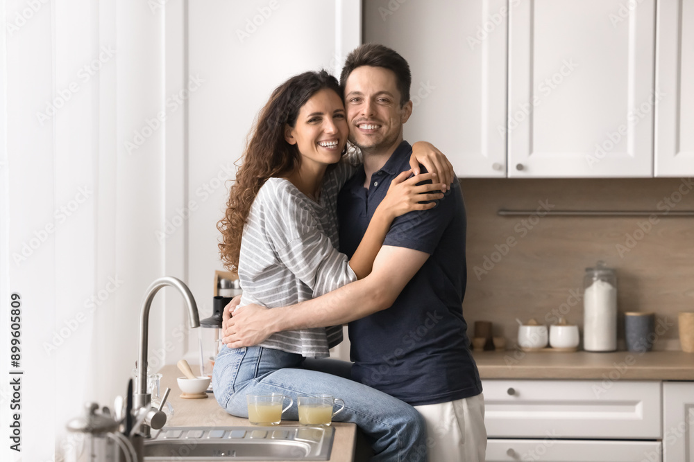 Beautiful married couple cuddling posing for camera in the kitchen. Happy Hispanic woman sits on countertop, hugs handsome smiling husband looking at cam. Homeowners family portrait, love, relations Beautiful married couple cuddling posing for camera in the kitchen. Happy Hispanic woman sits on countertop, hugs handsome smiling husband looking at cam. Homeowners family portrait, love, relations
