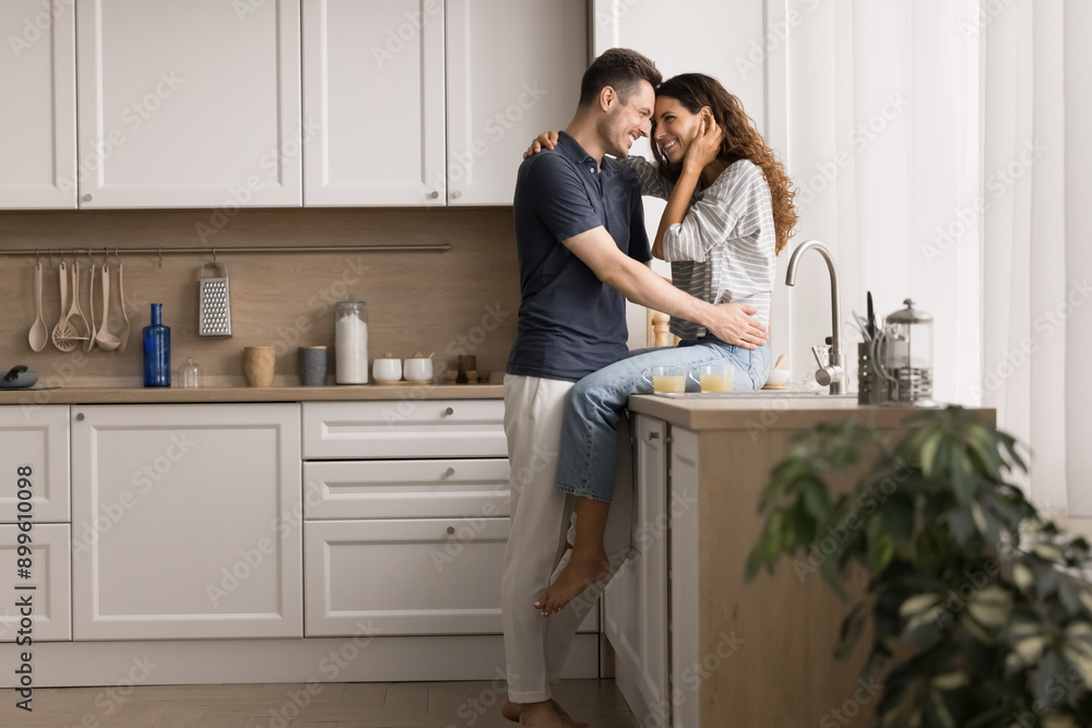 Young married couple sharing tender moment in modern kitchen. Pretty woman sitting on counter while her husband man stands close to her, gently hugging and looking into her eyes with love and warmth Young married couple sharing tender moment in modern kitchen. Pretty woman sitting on counter while her husband man stands close to her, gently hugging and looking into her eyes with love and warmth
