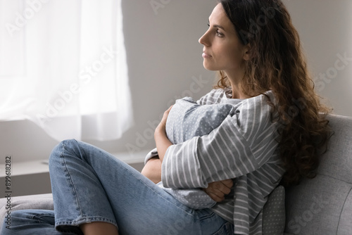 Pensive single woman sits on sofa, gazing out of window in deep contemplation, suffers from solitude, emotional distress, coping with personal troubles and break up, goes through difficult life period