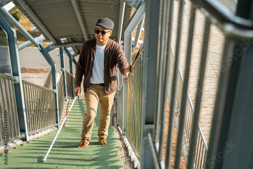 Young blind man walking in city with walking stick. Visually impaired man difficult to traveling on the road wearing sun glasses Cross the road cross the footbridge © Chanakon