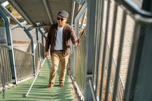 Young blind man walking in city with walking stick. Visually impaired man difficult to traveling on the road wearing sun glasses Cross the road cross the footbridge photo