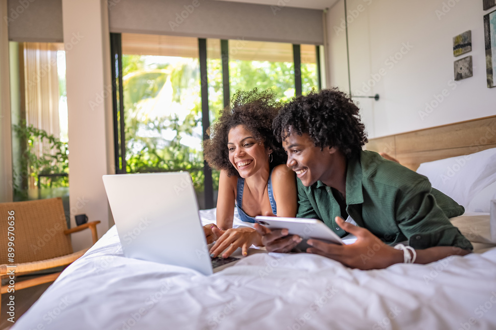 Fototapeta premium A young couple of African descent is lying on a bed in a modern, sunlit bedroom, smiling and looking at a laptop screen. They appear to be enjoying a pleasant moment together, relaxed and happy.