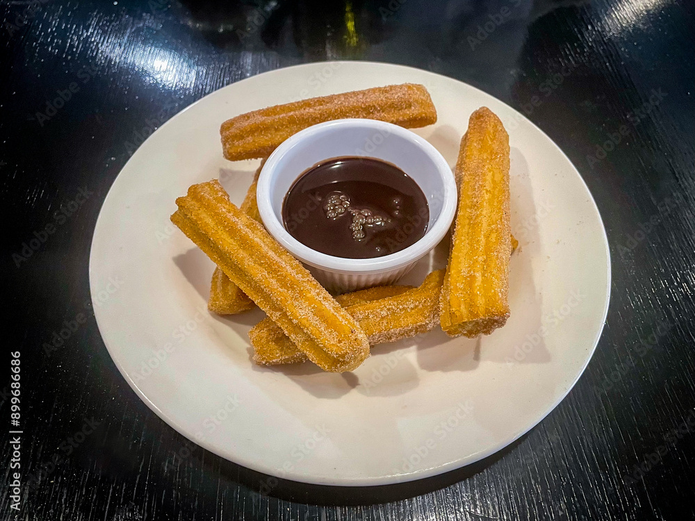 A plate of golden-brown churros dusted with sugar is served with a small ramekin of rich, dark chocolate sauce. The churros are crispy and perfectly shaped, inviting a delightful and indulgent treat.
