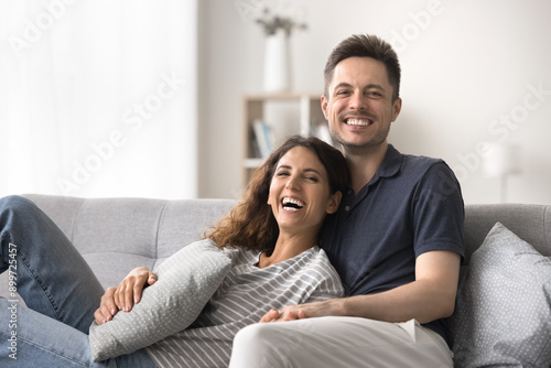 Portrait of happy millennial boyfriend and girlfriend looking at camera laughing, relaxing on comfortable couch at home, spend pastime together. Strong marriage, harmonic relationships, understanding