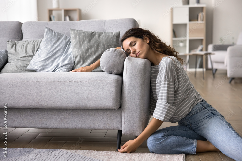 Young fatigued, overworked woman leaning against sofa, rests after hard work, fell asleep on floor in living room, her posture and expression convey a deep sense of physical and emotional tiredness Young fatigued, overworked woman leaning against sofa, rests after hard work, fell asleep on floor in living room, her posture and expression convey a deep sense of physical and emotional tiredness