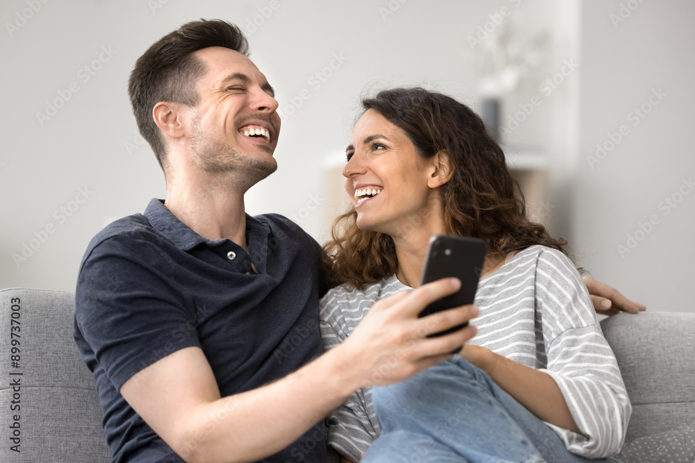 Obraz premium Cheerful young wife showing funny videos on social media network on cellphone to laughing husband, sit together on sofa, resting on weekend using new mobile app, giggling, having fun. Tech, leisure