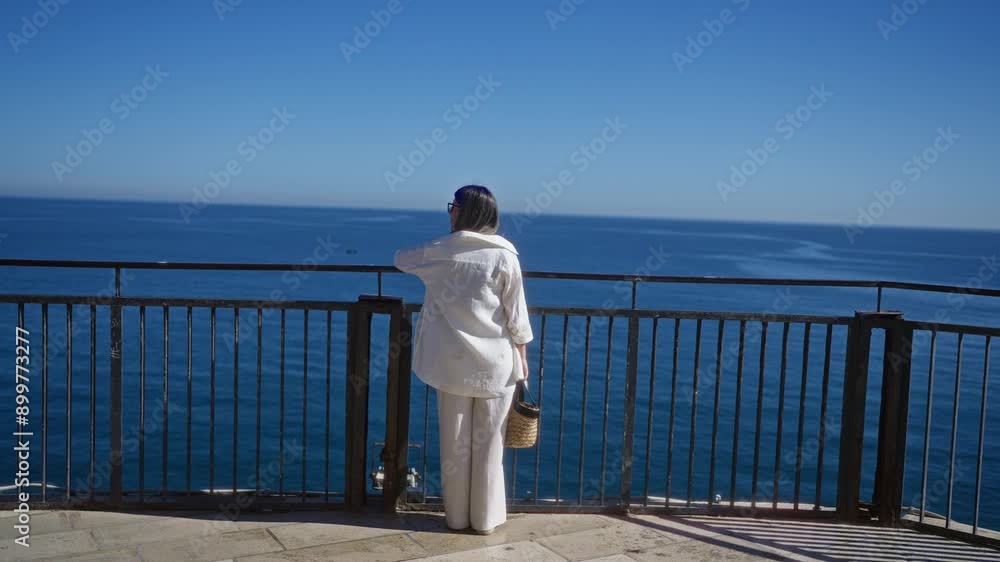 A young hispanic woman in white attire stands at a viewpoint overlooking the serene blue sea at polignano a mare, puglia, italy on a clear, sunny day. A young hispanic woman in white attire stands at a viewpoint overlooking the serene blue sea at polignano a mare, puglia, italy on a clear, sunny day.