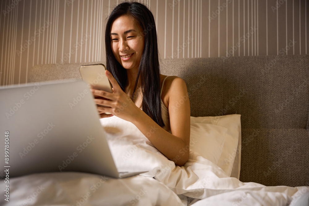 Smiling Asian woman texting on smartphone and laptop sitting in bed Smiling Asian woman texting on smartphone and laptop sitting in bed