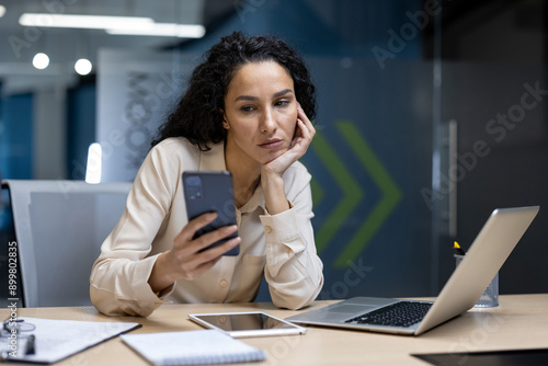 Bored businesswoman sitting at desk, staring at smartphone. Office environment with laptop, tablet, and notepad on desk. Concept of workplace boredom, lack of motivation, or distraction
