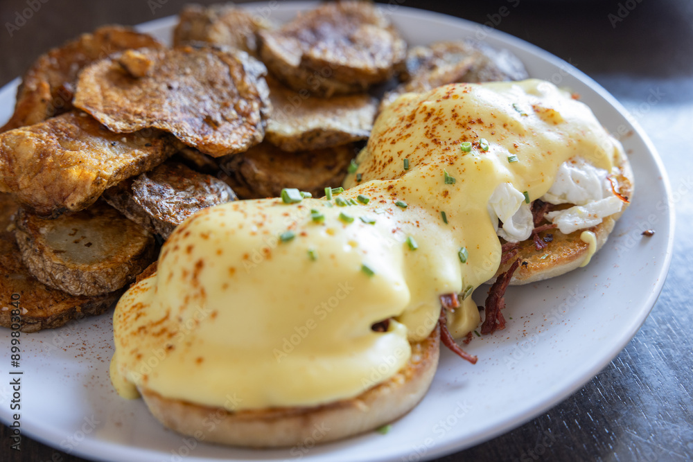 Eggs Benedict served with extra sauce and sliced potatoes on a white plate and garnished with paprika and chives. Eggs Benedict served with extra sauce and sliced potatoes on a white plate and garnished with paprika and chives.