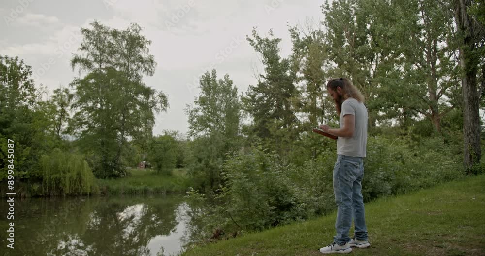 Young man stands by a lake in a scenic forest, using his tablet on a tranquil day. The serene environment features lush greenery and still waters. Young man stands by a lake in a scenic forest, using his tablet on a tranquil day. The serene environment features lush greenery and still waters.