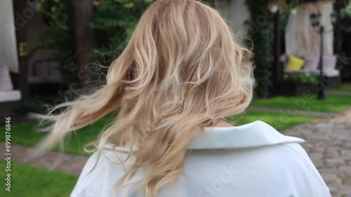 A blonde girl walks along the alley, filmed from behind, from the back. White curls blow in the wind