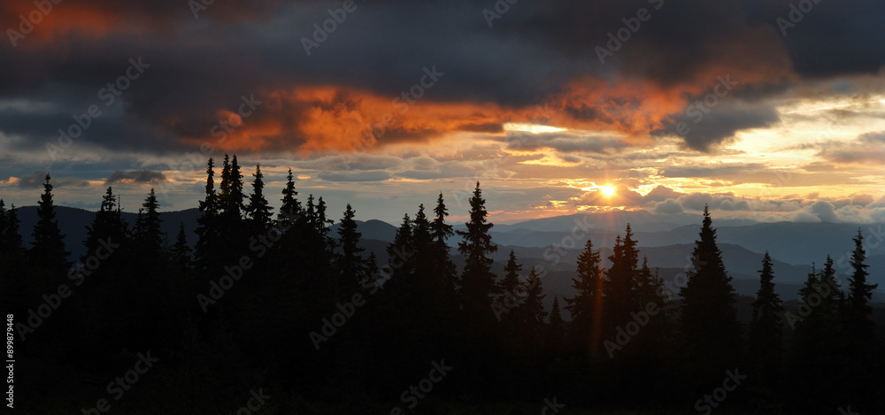 Dramatic sunset paints sky with fiery orange and dark clouds, casting serene glow over dense forest of silhouetted pine trees. Distant mountain ridges add depth to this breathtaking natural landscape. Dramatic sunset paints sky with fiery orange and dark clouds, casting serene glow over dense forest of silhouetted pine trees. Distant mountain ridges add depth to this breathtaking natural landscape.