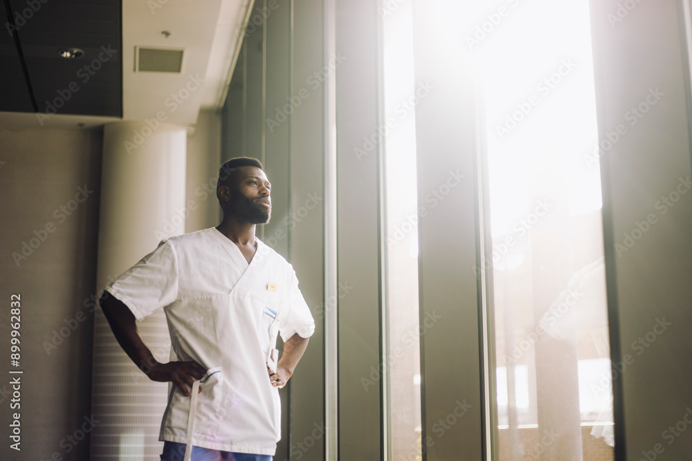 Low angle view of male medical professional standing with arms akimbo near window in hospital Low angle view of male medical professional standing with arms akimbo near window in hospital
