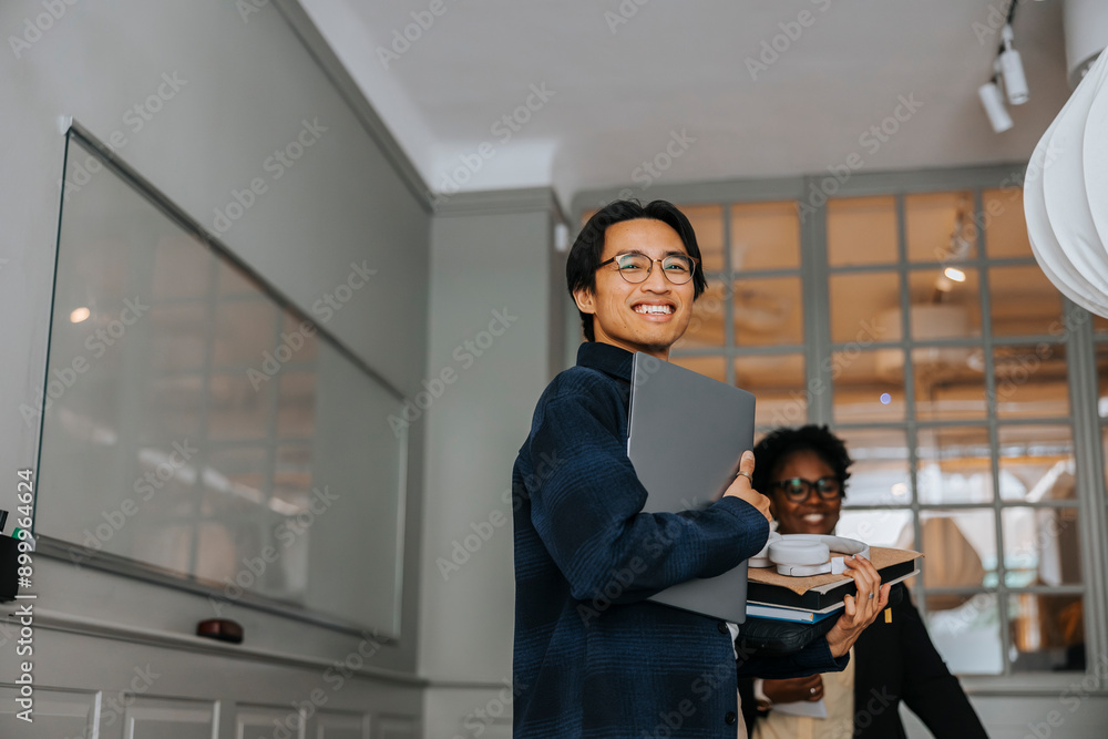 Happy male entrepreneur holding laptop while standing in meeting room Happy male entrepreneur holding laptop while standing in meeting room