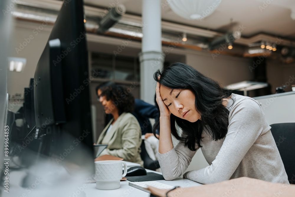 Exhausted female business professional with eyes closed sitting with head in hand near desk at office Exhausted female business professional with eyes closed sitting with head in hand near desk at office