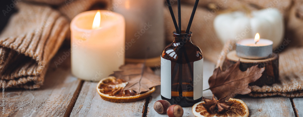 Fototapeta premium Hello autumn, cozy slow living. Pumpkin, aromatic candle, warm sweaters, dry fall leaves, cinnamon, anise in golden lights with bokeh on white wooden background. Thanksgiving apartment decor banner