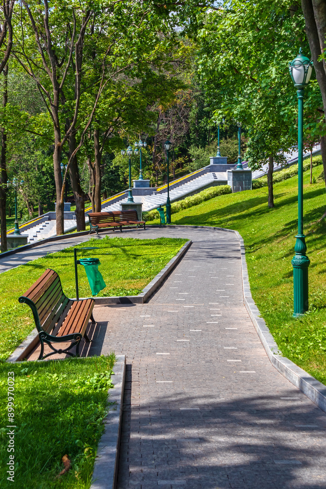 An alley with benches and a stone staircase on a slope in a park on a sunny summer day. Landscape of an urban garden with green lawns and vintage street lamps An alley with benches and a stone staircase on a slope in a park on a sunny summer day. Landscape of an urban garden with green lawns and vintage street lamps