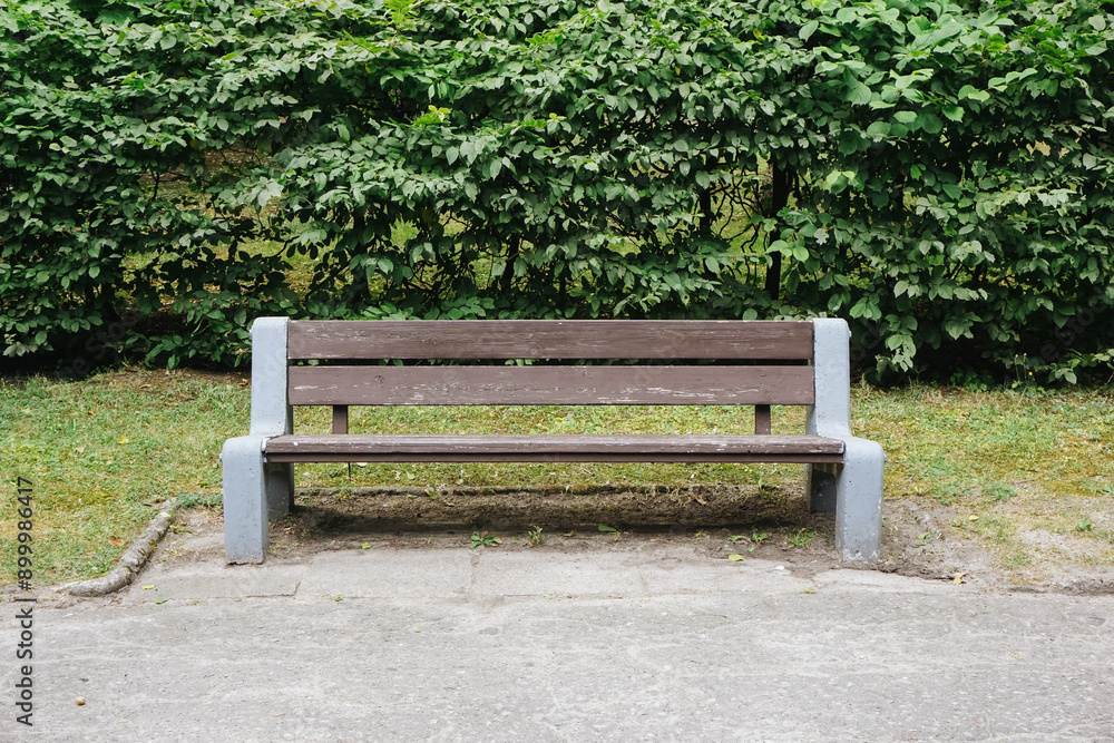 Wooden and concrete bench in a park. Empty bench by the pathway with lawn and bushes in the background. Tranquil outdoor setting ideal for relaxation and enjoying nature. Simple city park landscape. Wooden and concrete bench in a park. Empty bench by the pathway with lawn and bushes in the background. Tranquil outdoor setting ideal for relaxation and enjoying nature. Simple city park landscape.